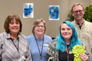 Photo of four people at the IMAGES Artist Dinner and Award Ceremony where the artist, who has bright blue hair and is holding a green ribbon, has won an award.